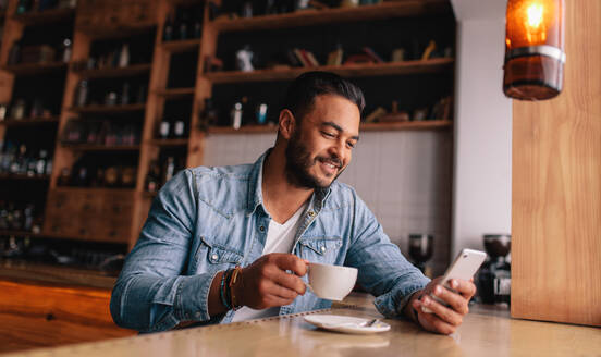 Hübscher junger kaukasischer Mann, der in einem Café sitzt, sein Handy benutzt und Kaffee trinkt. Kaukasischer Mann mit einer Tasse Kaffee und einer Textnachricht auf seinem Smartphone. - JLPSF26883