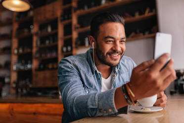 Handsome young man sitting at cafe making video call from his mobile phone. Caucasian male at coffee shop having a videochat on smart phone. - JLPSF26886