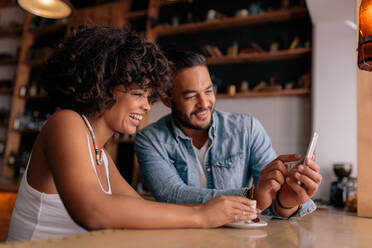 Junges glückliches Paar sitzt am Tisch in einem Café und schaut auf sein Handy und lächelt. Afrikanische Frau und kaukasischer Mann in einem Café und benutzt ein Smartphone. - JLPSF26889