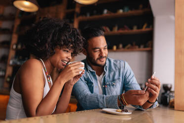 Afrikanische Frau, die mit ihrem Freund Kaffee trinkt und etwas auf dem Handy zeigt. Junges Paar im Café, das auf sein Smartphone schaut. - JLPSF26890