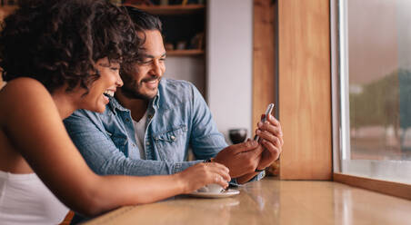 Lächelndes junges Paar, das in einem Café sitzt und auf sein Handy schaut. Junger Mann und Frau in einem Café, die ein Smartphone benutzen. - JLPSF26891