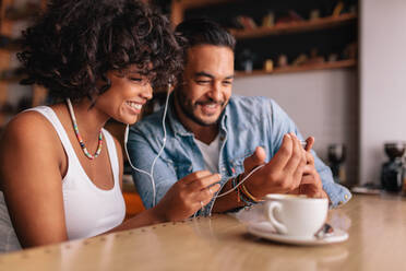 Happy young couple sitting at coffee shop having video chat on mobile phone. Young man and woman with earphones at cafe using smart phone. - JLPSF26892