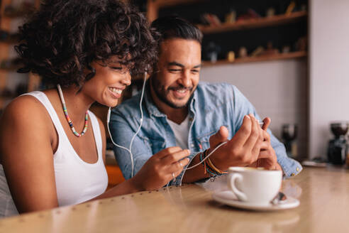 Glückliches junges Paar sitzt in einem Café und führt einen Videochat mit dem Handy. Junger Mann und Frau mit Kopfhörern in einem Café und benutzen ein Smartphone. - JLPSF26892