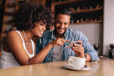 Lächelndes junges Paar mit Kopfhörern, das in einem Café sitzt und auf sein Mobiltelefon schaut. Junger Mann und Frau in einem Café, die ein Smartphone benutzen. - JLPSF26893