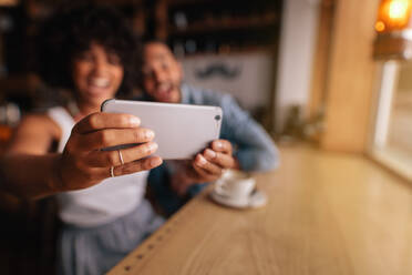 Junge Frau nimmt Selbstporträt mit Handy, während sie mit ihrem Freund in einem Café sitzt. Paar nimmt Selbstporträt mit Smartphone. Fokus auf Handy in den Händen der Frau. - JLPSF26898