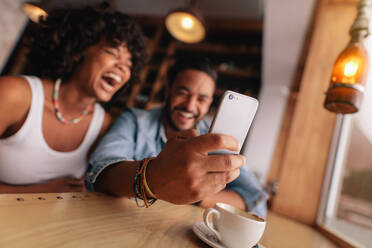 Junges Paar im Café, das ein Selfie mit dem Smartphone macht. Junger Mann und Frau lachen, während sie ein Selbstporträt im Café machen. - JLPSF26907