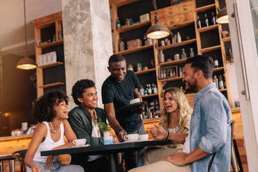 Eine Gruppe von Freunden sitzt am Tisch und der Kellner serviert Kaffee in einem Café. Junge Leute treffen sich in einem Café. - JLPSF26908