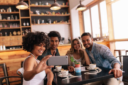 Multirassische Menschen, die sich in einem Café amüsieren und ein Selfie mit ihrem Mobiltelefon machen. Eine Gruppe junger Freunde sitzt in einem Restaurant und macht ein Selbstporträt mit ihrem Smartphone. - JLPSF26914