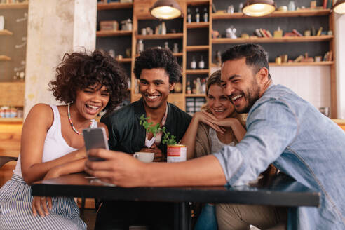 Eine Gruppe junger Leute sitzt in einem Café und schaut sich die Fotos auf ihrem Smartphone an. Junge Männer und Frauen sitzen am Cafétisch und benutzen ihr Handy. - JLPSF26916