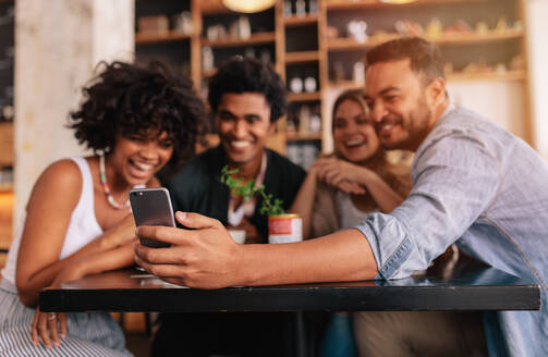 Junge Männer und Frauen sitzen am Cafétisch und nehmen ein Selbstporträt mit dem Handy auf. Multirassische Gruppe von Freunden, die ein Selfie mit dem Smartphone im Café machen. - JLPSF26917