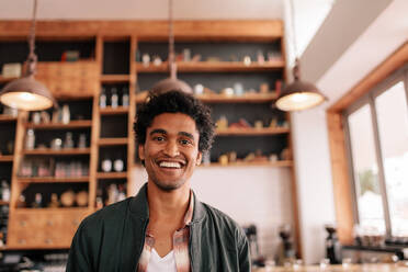 Portrait of happy young man standing in a cafe. Handsome young african guy standing in a coffee shop and smiling. - JLPSF26925