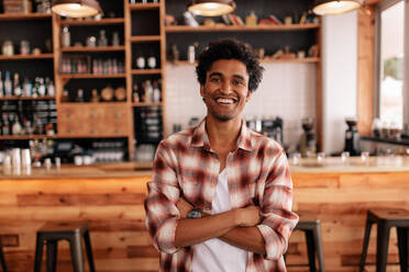 Porträt eines gutaussehenden jungen Mannes mit verschränkten Armen in einem Café. Lächelnder afrikanischer Mann, der in einem Café steht. - JLPSF26928