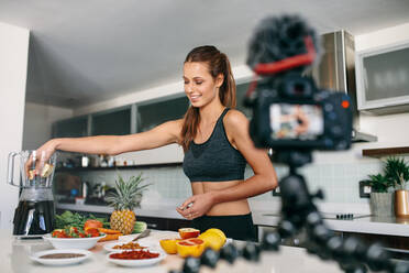 Young lady in sports wear preparing fruit juice while a tripod mounted camera is recording a video. Woman recording content for her vlog in kitchen. - JLPSF26937