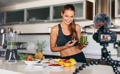 Smiling woman preparing a healthy breakfast with fruits and vegetables. Young lady holding glass of green juice at the kitchen table looking at a camera mounted on tripod. - JLPSF26941