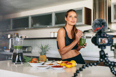 Young lady in kitchen drinking healthy vegetable juice while recording it on camera. Fruits and vegetables for breakfast on kitchen table. - JLPSF26943