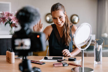 Frau, die ein Video für ihren Blog über Kosmetika dreht. Lächelnde junge Frau vor einer Aufnahmekamera mit verschiedenen Kosmetikartikeln auf dem Tisch. - JLPSF26944