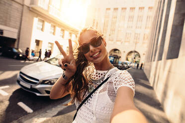 Tourist posiert für ein Selfie in einer Straße mit blinkendem Siegeszeichen. Vloggerin nimmt Inhalte für ihren Reise-Vlog auf. - JLPSF26956