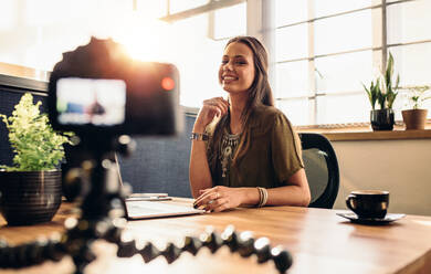 Junge Frau, die ein Video für ihren Vlog mit einer Digitalkamera auf einem flexiblen Stativ aufnimmt. Lächelnde Frau, die an ihrem Schreibtisch sitzt und an einem Laptop arbeitet. - JLPSF26965