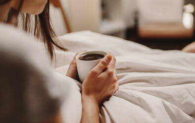 Nahaufnahme einer Frau auf dem Bett sitzend mit einer Tasse Kaffee in der Hand. Frau mit Kaffee in der Hand auf dem Bett sitzend. - JLPSF26972