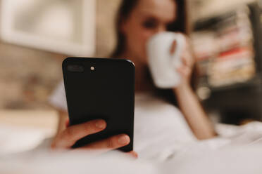 Frau schaut auf ihr Handy, während sie im Bett Kaffee trinkt. Frau sitzt auf dem Bett mit einer Tasse Kaffee und hält ein Handy in der Hand. - JLPSF26978