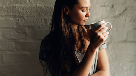 Nahaufnahme einer Frau, die mit einer Tasse Kaffee in der Hand an der Wand steht. Frau schaut auf die Kaffeetasse in ihrer Hand, die an einer Wand steht. - JLPSF26979