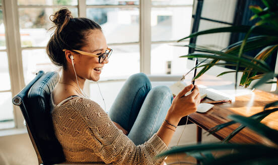 Seitenansicht einer lächelnden Frau, die auf ihr Mobiltelefon schaut und über Kopfhörer Musik hört. Eine Frau führt einen Videoanruf mit ihrem Mobiltelefon mit drahtloser Tastatur und Maus auf dem Tisch. - JLPSF26984