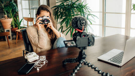 Frau, die mit einer Kamera fotografiert, während sie zu Hause sitzend Inhalte für ihren Blog aufnimmt. Bloggerin, die Inhalte für ihren Blog mit einer Kamera auf einem flexiblen Stativ und einem Laptop auf dem Tisch aufnimmt. - JLPSF26988