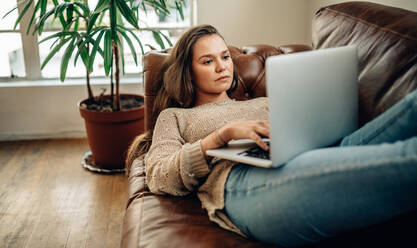 Frau, die zu Hause auf einem Sofa liegt und an einem Laptop-Computer arbeitet. - JLPSF27000