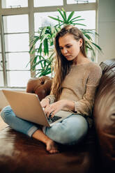 Frau sitzt mit gekreuzten Beinen auf einem Sofa zu Hause und arbeitet an einem Laptop-Computer. - JLPSF27001