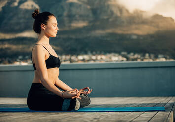 Frau, die auf der Terrasse ihres Hauses Yoga praktiziert. Fitness-Frau, die in einer Yoga-Haltung auf dem Boden sitzend mit gekreuzten Beinen meditiert. - JLPSF27007