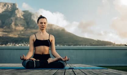 Eine Frau, die auf der Terrasse ihres Hauses Yoga praktiziert, meditiert in einer Yogahaltung, sitzt mit gekreuzten Beinen auf dem Boden und hört über Kopfhörer Musik. - JLPSF27008