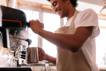 Professioneller Barista hält Metallkanne, die Milch in der Kaffeemaschine erwärmt. Glücklicher junger Mann bereitet Kaffee am Tresen zu. - JLPSF27013