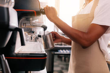 Nahaufnahme eines jungen Mannes, der an einer Maschine Kaffee zubereitet. Der Barista hält eine Metallkanne, in der er Milch erwärmt. - JLPSF27015
