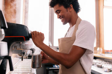 Ein lächelnder Barista hält eine Metallkanne in der Hand und wärmt Milch in der Kaffeemaschine auf. Ein glücklicher junger Mann bereitet am Tresen Kaffee zu. - JLPSF27016