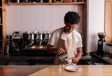 Junger afrikanischer Mann gießt Milch in Kaffee und macht Espresso. Professioneller Barista bereitet Kaffee auf dem Tresen zu. - JLPSF27017