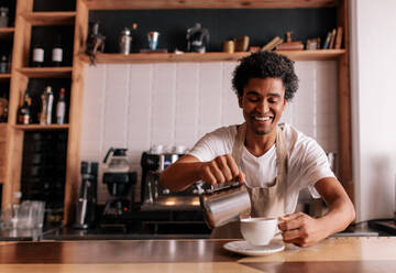 Professioneller Barista macht Kaffee auf dem Tresen eines Cafés. Junger afrikanischer Mann gießt Milch in den Bullen und lächelt. - JLPSF27018