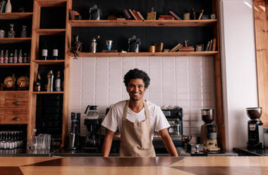 Portrait of professional barista standing at cafe counter. African man in apron looking at camera and smiling. - JLPSF27019