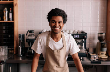 Porträt eines glücklichen jungen Barista, der an der Theke eines Cafés steht. Afrikanischer Mann in Schürze schaut in die Kamera und lächelt. - JLPSF27020