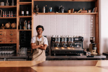 Portrait of confident young man wearing apron standing behind cafe counter. Barista in apron looking at camera and smiling. - JLPSF27021