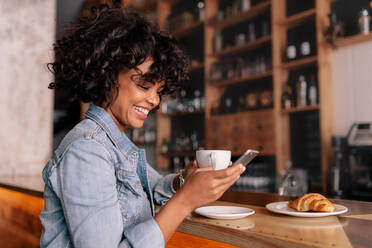 Lächelnde Frau, die ein Smartphone in einem modernen Café benutzt. Junge afrikanische Frau, die in einem Café sitzt, Kaffee trinkt und eine Textnachricht auf ihrem Handy liest. - JLPSF27038