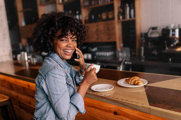 Lächelnde junge Frau, die in einem modernen Café mit ihrem Handy telefoniert. Lachende junge Afrikanerin, die in einem Café sitzt, einen Kaffee trinkt und ein Telefonat führt. - JLPSF27039