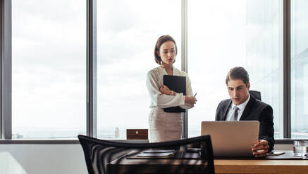 Zwei Geschäftsleute, die auf einen Laptop schauen, während sie im Büro arbeiten. Ein Geschäftsmann sitzt an seinem Schreibtisch und eine Mitarbeiterin steht daneben. - JLPSF27042