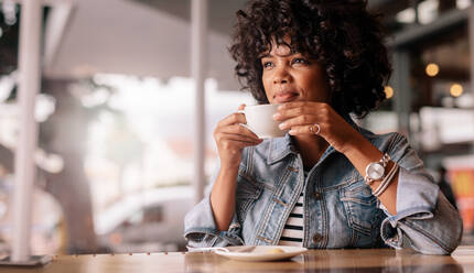 Porträt einer jungen afrikanischen Frau, die in einem Café sitzt und eine Tasse Kaffee in der Hand hält, wegschaut und nachdenkt. Nachdenkliche junge Frau beim Kaffee trinken in einem Restaurant. - JLPSF27044