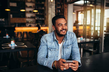Innenaufnahme eines jungen Mannes, der mit seinem Handy in einem Café sitzt. Gut aussehender kaukasischer Mann im Café, der wegschaut und lächelt. - JLPSF27045