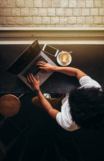 Draufsicht auf einen jungen Mann, der in einem Café sitzt und an einem Laptop arbeitet. Vertikale Draufsicht auf einen Mann, der in einem Café einen Laptop benutzt. - JLPSF27046