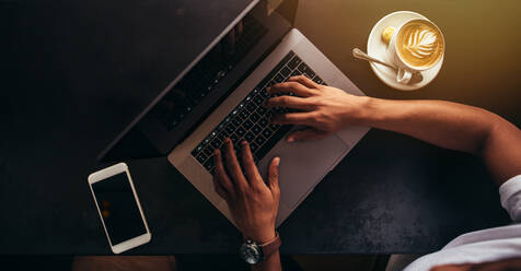 Close up shot of young man sitting at coffee shop and working on laptop. Focus on hands typing on laptop keyboard with mobile phone and cup of coffee on table. - JLPSF27048