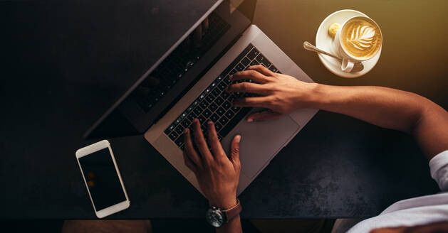 Nahaufnahme eines jungen Mannes, der in einem Café sitzt und an einem Laptop arbeitet. Fokus auf die Hände, die auf der Tastatur des Laptops tippen, mit einem Mobiltelefon und einer Tasse Kaffee auf dem Tisch. - JLPSF27048