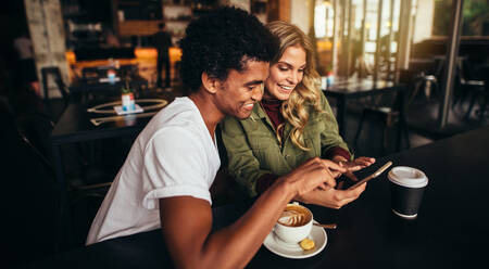 Aufnahme eines glücklichen jungen Mannes und seiner Freundin, die zusammen in einem Café sitzen und ihr Handy benutzen. Beste Freunde in einem Café, die auf ihr Smartphone schauen. - JLPSF27049