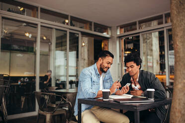 Zwei junge Männer sitzen zusammen in einem Café und benutzen ein Mobiltelefon. - JLPSF27052