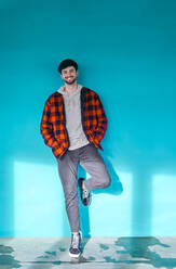 Glücklicher Mann mit Bart in Freizeitkleidung schaut in die Kamera, während er in der Nähe einer blauen Wand in einem hellen geräumigen Gebäude mit Fenstern steht - ADSF39934